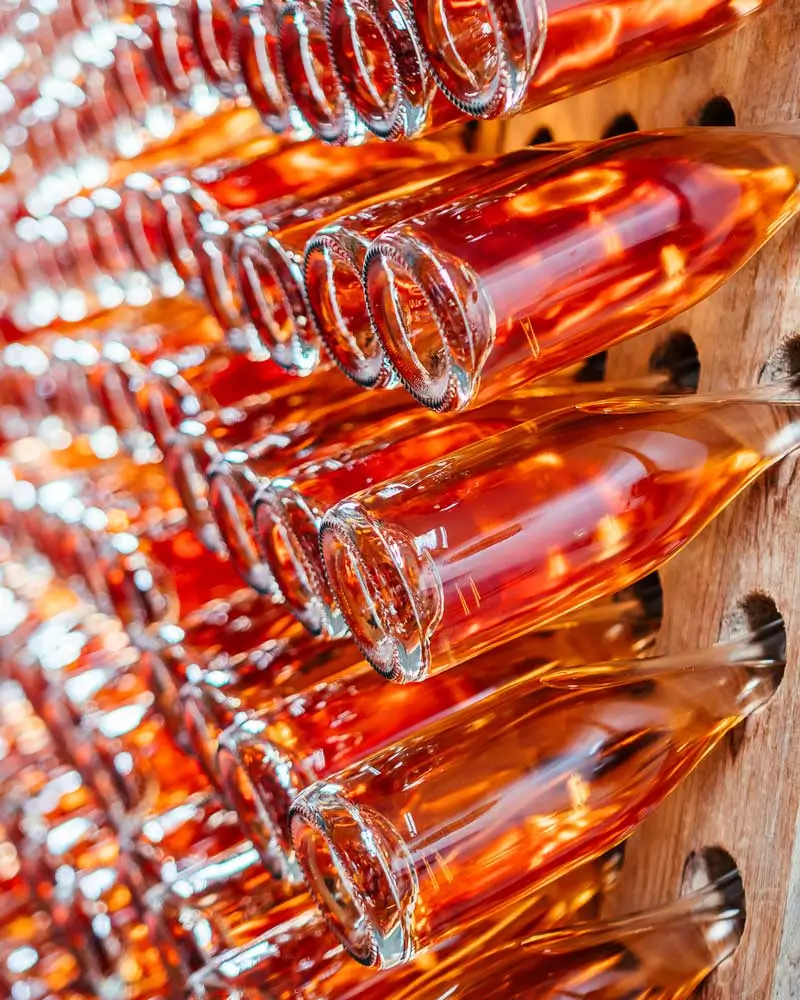 Close-up of rows of amber-hued wine bottles resting neck-down in a wooden riddling rack. Warm light illuminates the glass bottoms and the glowing liquid inside, creating a repetitive, textured pattern.